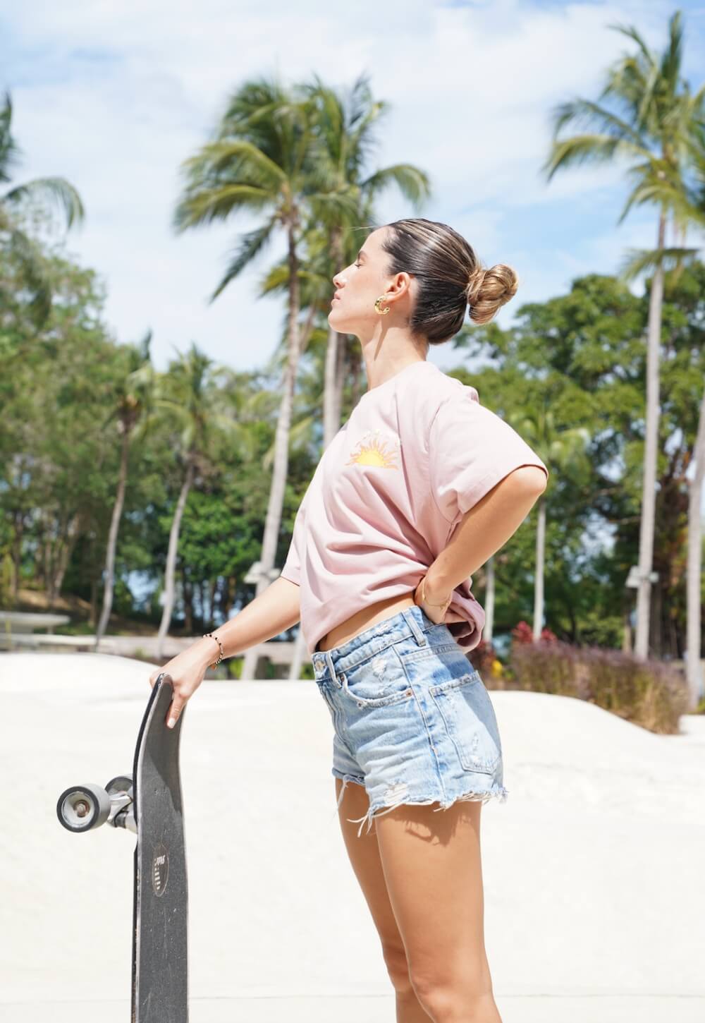 Woman wearing a pink Salty t-shirt and denim shorts holding a skateboard in a tropical skate park